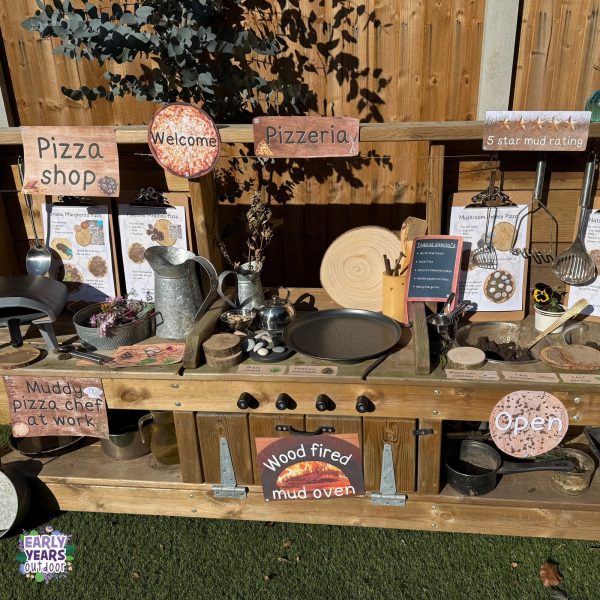 Pizza shop role play signs set up in the mud kitchen with pizza mud kitchen recipes, bowls of pinecones, mud, utensils, jugs, pizza trays, pebbles and other nature